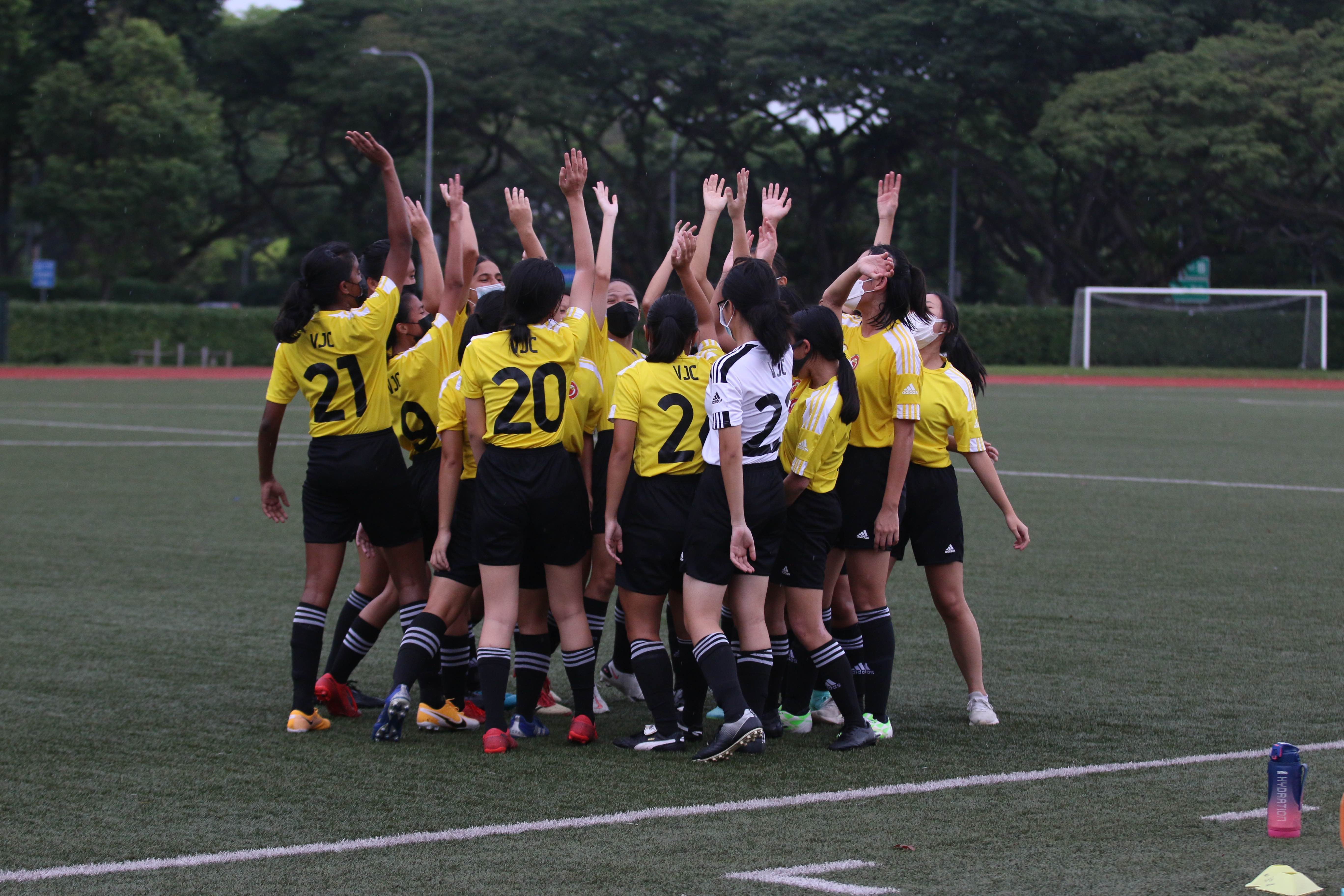 A girls soccer team in yellow VJC jerseys celebrates on a green field with hands raised.